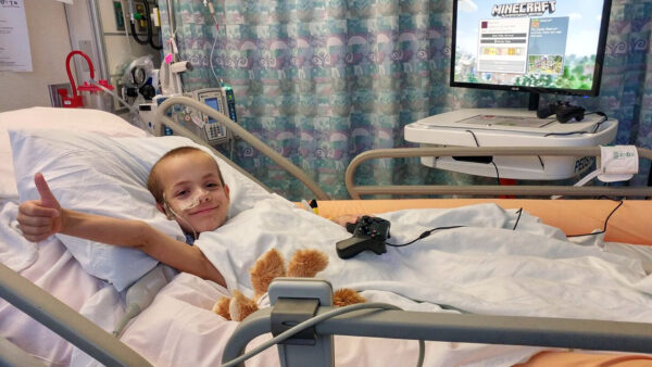 Kid sitting on a hospital bed, smiling, while looking at the camera and giving a thumbs up. He is playing Minecraft on a GO Kart.